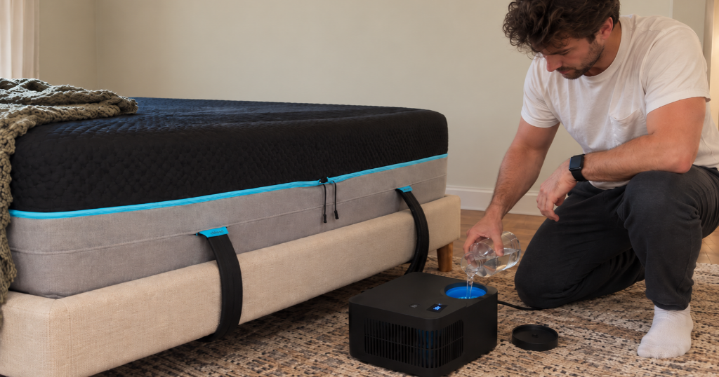 Man refilling the Chilipad 2.0 Dock with distilled water beside a bed fitted with the cooling system.